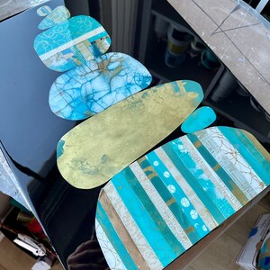 Puede incluir: Pieza de arte abstracto con una pila de piedras superpuestas, texturizadas y pintadas en tonos azules, dorados y blancos sobre un fondo negro.