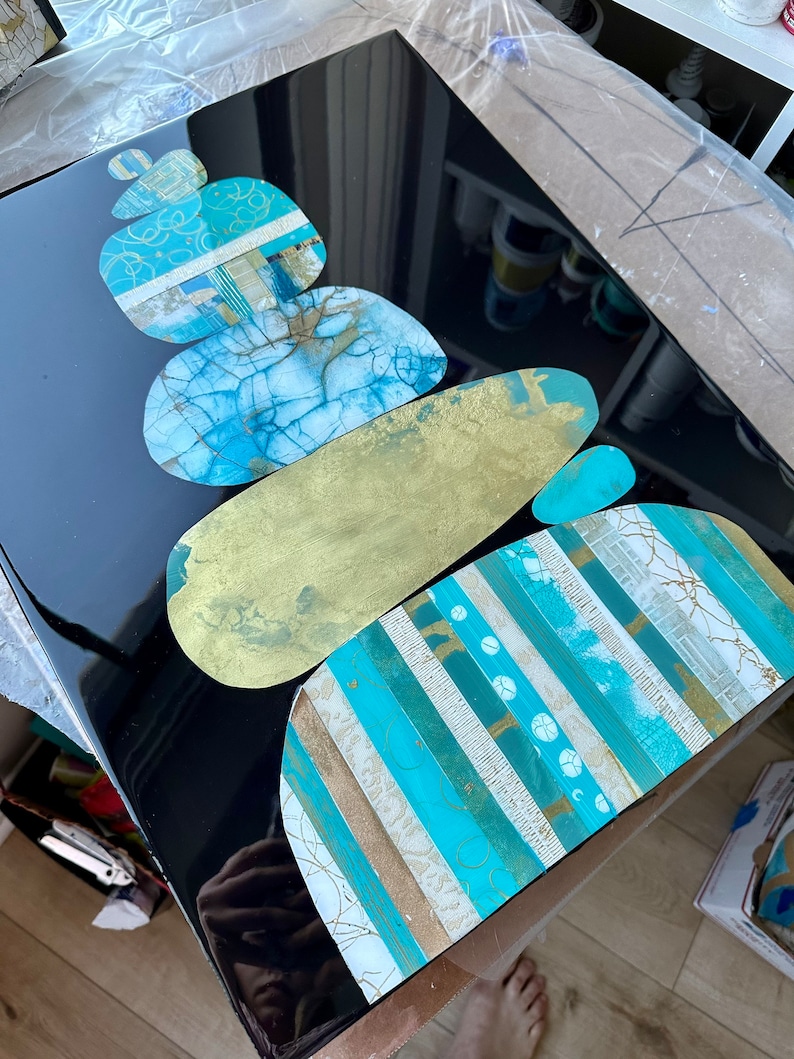 Puede incluir: Pieza de arte abstracto con una pila de piedras superpuestas, texturizadas y pintadas en tonos azules, dorados y blancos sobre un fondo negro.