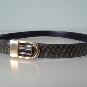 May include: A grey snakeskin belt with a gold-toned buckle. The buckle has a black rectangular insert.
