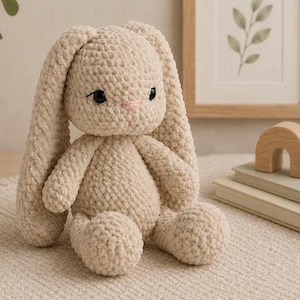 Puede incluir: Un conejo de peluche tejido a ganchillo en color beige con orejas largas y caídas, acabado de hilo suave y texturizado, pequeños ojos negros de seguridad y una nariz delicadamente bordada en rosa. El peluche está sentado con extremidades.