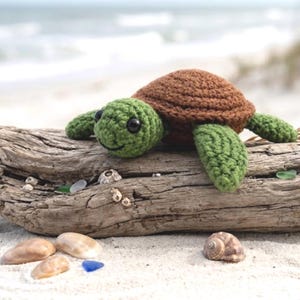 May include: A crocheted turtle toy featuring a textured brown shell and a green head with black plastic eyes. The turtle has four green flippers and a small stitched mouth. The yarn construction shows visible loops throughout the body and shell.