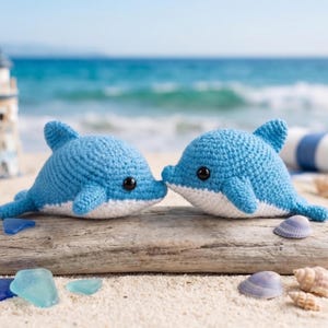 May include: Two blue crochet dolphins with white bellies are touching noses on a piece of driftwood. The dolphins have black eyes and are set against a beach scene with seashells and the ocean in the background.