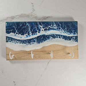 May include: Wooden key holder with a resin ocean wave design. The top section features a deep blue and white wave pattern, transitioning to a sandy beach design. Four white metal hooks are attached to the wooden base for hanging keys.