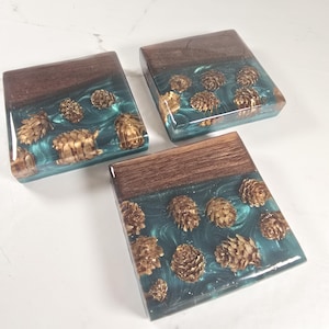 May include: Three square coasters with a dark wood top and a teal resin base. The resin is embedded with small, golden pinecones. The coasters are arranged on a white surface.