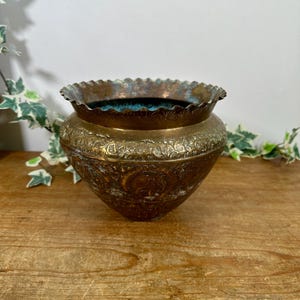 May include: A round brass bowl featuring a scalloped rim and intricate embossed floral and animal motifs across its exterior surface. The metal displays a weathered patina with dark brown and oxidised green tones, and the interior is a deep teal.