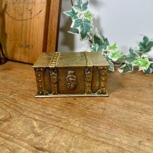 May include: A rectangular brass box designed to resemble a miniature wooden chest. The exterior features a textured wood grain pattern, reinforced brass-coloured corner accents, and a central crest depicting a lion emblem on the front panel.