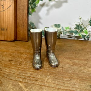 May include: A pair of miniature metallic silver riding boots with a smooth, reflective finish and a white interior lining. The boots feature a tall, structured shaft, a rounded toe, and a small heel, standing upright as a decorative object.