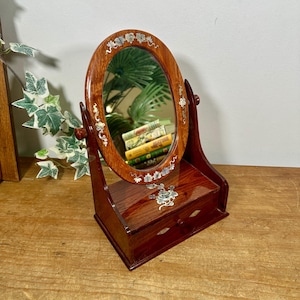 May include: A polished wooden vanity mirror with an oval frame and a base featuring a single drawer. The frame and drawer front are decorated with inlaid mother-of-pearl floral and vine motifs. The mirror pivots on two side supports.