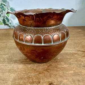 May include: A copper planter with a wide, scalloped rim and a rounded body. The upper section features a textured, hammered finish, while the middle band displays a series of embossed, semi-circular arches with subtle green patina accents.
