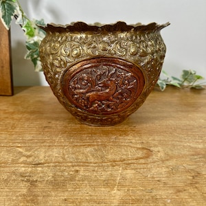 May include: A brass bowl featuring an intricate embossed floral pattern across its surface. The centre of the bowl displays a prominent oval medallion with a raised relief depiction of a deer surrounded by detailed foliage and scrollwork.
