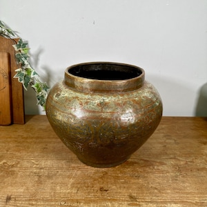 May include: A round, wide-mouthed metal vessel with a dark, weathered copper and brass finish. The exterior surface is decorated with intricate, engraved patterns and repeating motifs that wrap around the body of the container.