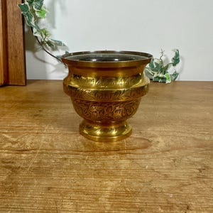 May include: A polished brass bowl featuring a wide, flared rim and a pedestal base. The exterior surface is decorated with intricate, engraved leaf patterns that wrap around the midsection and the base of the vessel, creating a textured appearance.