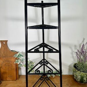May include: Black metal corner shelf with three triangular shelves. The shelves are supported by a decorative wrought iron frame. The shelf is designed to fit into a corner and is suitable for displaying decorative items or plants.