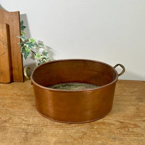 May include: A round copper basin with a polished metallic finish and two brass handles attached to the sides. The interior of the basin shows a weathered, greenish patina, while the exterior surface displays a warm, reddish-brown copper tone.
