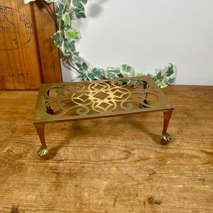 May include: A rectangular brass trivet with ornate cut-out designs and four small, claw-footed legs. The trivet is a warm gold colour and sits on a wooden surface. Decorative ivy and a wooden cheese board are in the background.