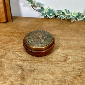 May include: A round wooden box featuring a dark brown grain and a circular lid. The lid is made of a metallic material with a weathered, greenish-bronze patina and an intricate, embossed relief pattern depicting floral and foliage motifs.