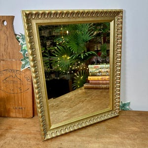 May include: A rectangular wall mirror featuring a decorative gold-toned frame with a repeating scalloped or wave-like pattern along the outer edge. The mirror glass is clear and reflects a stack of books, a small figurine, and green foliage.