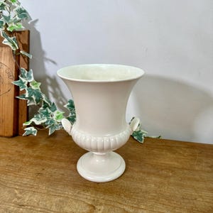 May include: A cream-coloured ceramic urn-style vase featuring a wide, flared rim, a ribbed lower body, and two small decorative handles on the sides. The vase stands on a circular pedestal base and has a smooth, glossy finish throughout its exterior.