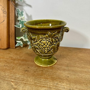 May include: A moss green ceramic planter with a pedestal base and two small side handles. The exterior features a raised, embossed relief pattern of grape clusters and vine leaves encircling the body of the vessel.
