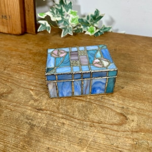 May include: A rectangular stained glass box featuring a geometric pattern of blue, light blue, and purple glass panels. The lid is decorated with two pink rose motifs set against a blue background, all held together by thin silver-toned metal solder.