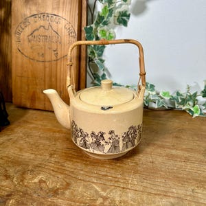May include: A cream ceramic teapot with a bamboo handle and lid. The teapot has a decorative band of black and white figures around the base. It sits on a wooden surface, with a wooden sign in the background.