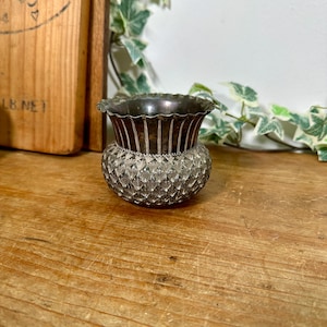 May include: A small, round, silver-toned metal vessel featuring a scalloped, flared rim and a textured body with a repeating diamond-shaped pattern. The upper section of the vessel has vertical fluting, while the base is rounded and bulbous.
