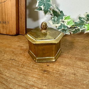 May include: A hexagonal brass box with a matching lid featuring a central knob handle. The container has a polished metallic finish with visible signs of age and patina, showcasing a six-sided geometric shape with a flat base and angled sides.