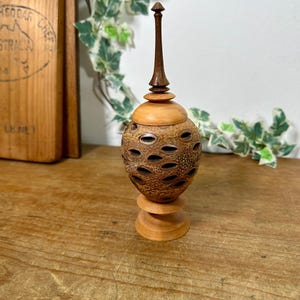 May include: A handcrafted wooden decorative vase with a unique, textured design. The vase features a light brown base and lid, with a dark brown, elongated finial. The body has a series of oval openings. The vase stands approximately 15 cm tall.