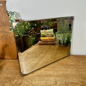 May include: Angled vintage mirror with metal corner brackets. The mirror reflects a wooden surface, books, and a green ceramic pot with a plant. A wooden cutting board is visible on the left.