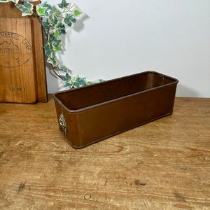 May include: A rectangular, copper-coloured loaf tin with decorative handles. The baking tin is made of metal and sits on a wooden surface. A wooden chopping board and greenery are in the background.