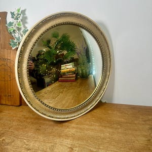 May include: A round wall mirror featuring a textured gold-toned frame with a beaded inner edge. The reflective surface shows a room interior with a stack of books, a small potted plant, and a green leafy plant, all captured in the mirror's reflection.