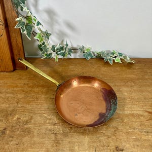 May include: A round copper frying pan with a long, slender brass handle. The interior of the pan shows a warm metallic copper surface with a distinct area of green oxidation and dark discolouration along the right side of the basin.