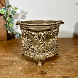 May include: A cylindrical olive-green ceramic planter featuring a raised relief design of figures and foliage. The vessel has a wide rim, a textured border, and stands on three decorative feet shaped like shells, providing a stable base.