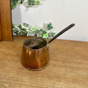 May include: A small copper Turkish coffee pot with a dark, slender metal handle. The pot features a polished, rounded body with a distinct pouring spout and a weathered interior surface, showcasing a classic design for brewing coffee.