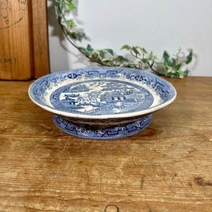 May include: A blue and white ceramic cake stand featuring a Willow pattern design. The stand has a round base and a matching plate top, ideal for displaying cakes or small items. The stand is on a wooden surface.