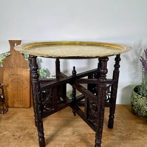 May include: A round brass tray with an intricate engraved pattern resting on a dark brown wooden folding base. The base features four carved legs connected by a cross-frame structure with detailed relief carvings on the side panels.