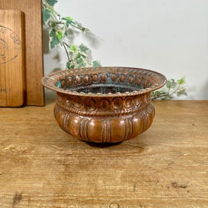 May include: A round copper bowl featuring a wide, flared rim with a repeating circular embossed pattern. The exterior body of the bowl has vertical fluted detailing, and the metal surface shows signs of oxidation with visible green patina.