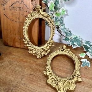 May include: Two ornate, gold-coloured oval picture frames. Each frame features cherubic figures and floral details. The frames are displayed on a wooden surface, with a wooden box in the background. Decorative ivy with green and white leaves is also present.