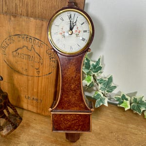 May include: A wall-mounted clock with a dark brown burl wood finish and a circular face. The clock face features black Roman numerals, gold-tone hands, and a central floral motif with small orange and green flowers. A gold-tone bezel surrounds the dial.