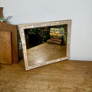 May include: A rectangular wall mirror featuring a frame with a light-coloured floral pattern in shades of beige, pink, and green. The mirror surface reflects a stack of books and green foliage, showing a clear glass pane within the decorative border.