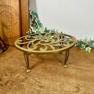 May include: An oval brass trivet featuring an intricate openwork scroll pattern on the top surface. The metal stand is supported by four curved legs that elevate the platform, providing a stable base for hot cookware or decorative items.