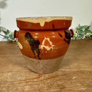 May include: A rustic ceramic pot with a wide opening and two small handles. The pot is glazed in a reddish-brown hue, adorned with dark brown and cream-coloured abstract patterns. The base has a lighter, unglazed finish, giving it a vintage look.