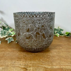 May include: A cylindrical metal planter featuring an intricate etched pattern of swirling foliage, birds, and a central human figure. The surface has a dark bronze finish with white detailing in the grooves and a scalloped, perforated top edge.