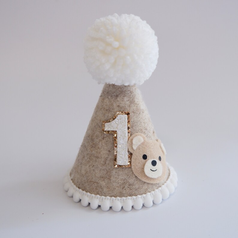 May include: A beige felt party hat with a white pom-pom on top. The hat has a white number "1" with gold glitter and a brown felt bear face on the front. The hat has a white pom-pom trim around the bottom.