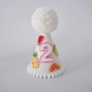 May include: White felt party hat with a white pom-pom on top. The hat has a pink glitter number "2" and colourful fruit appliques.