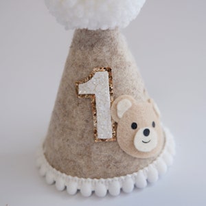 May include: A beige felt party hat with a white pom-pom on top. The hat has a gold glitter number "1" and a brown felt bear face on the front.