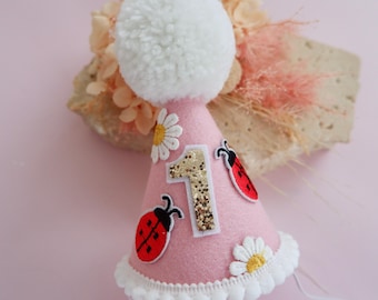 May include: A pink party hat with a white pom-pom on top, decorated with a gold glitter number 1, ladybug and daisy appliqués. The hat has a white pom-pom trim and a white string. Perfect for a first birthday celebration.