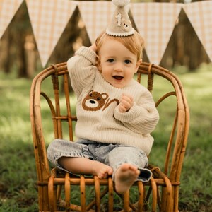 May include: A baby wearing a cream sweater with a brown bear applique and the number 'one' on it, sits in a wicker chair. The baby is wearing a brown felt party hat with a white pom-pom on top and the number 'one' on the front. The baby is smiling and looking at the camera.