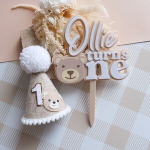 May include: A beige and white cake topper with the text "Ollie turns one" and a brown bear face. A beige felt party hat with a white pom-pom and a brown bear face is also included.
