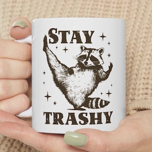 Può includere: Tazza in ceramica bianca con un'illustrazione in bianco e nero di un procione che fa una posa di yoga. Il testo "STAY TRASHY" è stampato sulla tazza.